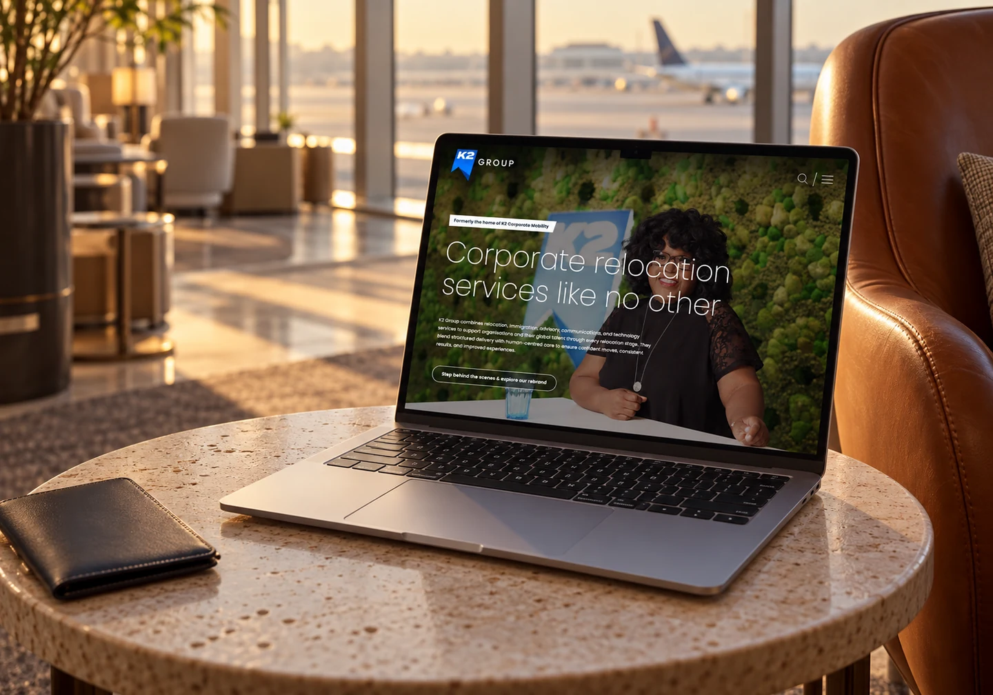 K2 Group homepage on a MacBook in a sunlit airport lounge.